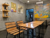 A cozy cafe interior with wooden tables and chairs. The walls are decorated with abstract paintings featuring vibrant and swirling patterns. A small wooden shelf on the left holds a decor item, possibly a miniature bicycle. Soft lighting creates a warm atmosphere.