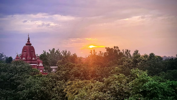 A picturesque landscape featuring a red temple with intricate architecture surrounded by lush green trees. The sun is setting in the horizon, casting a warm glow over the scene. The sky is partially cloudy, adding depth and texture to the image.