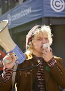 A person with light curly hair and freckles is speaking into a megaphone. The individual is wearing a brown jacket, a striped shirt, and a beanie adorned with the word 'SOLIDARITY'. The scene occurs outdoors in front of a building with the name 'CENTER COURT' visible in the background.