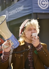 A person with light curly hair and freckles is speaking into a megaphone. The individual is wearing a brown jacket, a striped shirt, and a beanie adorned with the word 'SOLIDARITY'. The scene occurs outdoors in front of a building with the name 'CENTER COURT' visible in the background.