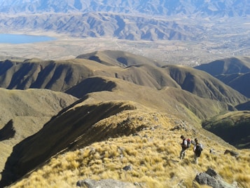 A vast mountainous landscape with rolling hills covered in grass and sparse vegetation. Two hikers with backpacks are walking along a ridge, capturing a scenic view of the terrain. A distant lake and sprawling valley can be seen below, surrounded by more rugged mountains in the background.