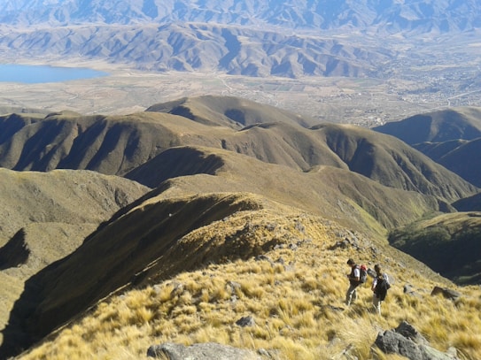 A vast mountainous landscape with rolling hills covered in grass and sparse vegetation. Two hikers with backpacks are walking along a ridge, capturing a scenic view of the terrain. A distant lake and sprawling valley can be seen below, surrounded by more rugged mountains in the background.