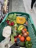 A shopping cart filled with a variety of fresh vegetables and fruits. Items visible include a halved pumpkin, radishes, bananas, tomatoes, onions, cauliflower, asparagus, avocado, and some leafy greens. The cart is in a grocery aisle, with the floor made of textured tiles and other shoppers passing by.