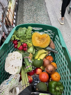 A shopping cart filled with a variety of fresh vegetables and fruits. Items visible include a halved pumpkin, radishes, bananas, tomatoes, onions, cauliflower, asparagus, avocado, and some leafy greens. The cart is in a grocery aisle, with the floor made of textured tiles and other shoppers passing by.