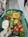 A shopping cart filled with a variety of fresh vegetables and fruits. Items visible include a halved pumpkin, radishes, bananas, tomatoes, onions, cauliflower, asparagus, avocado, and some leafy greens. The cart is in a grocery aisle, with the floor made of textured tiles and other shoppers passing by.