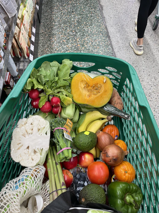 A close-up of a hand holding a shopping list next to a grocery cart filled with colorful produce.