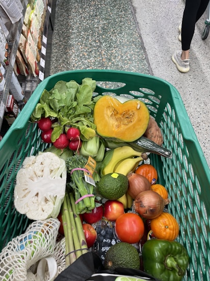 A shopping cart filled with a variety of fresh vegetables and fruits. Items visible include a halved pumpkin, radishes, bananas, tomatoes, onions, cauliflower, asparagus, avocado, and some leafy greens. The cart is in a grocery aisle, with the floor made of textured tiles and other shoppers passing by.