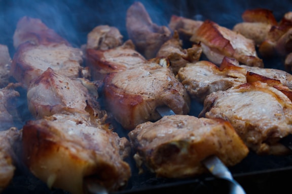 Juicy pieces of marinated meat are skewered and being grilled over an open flame. The meat appears to be evenly cooked with a crispy, golden-brown exterior, emanating a mouth-watering aroma accompanied by small wisps of smoke rising up.