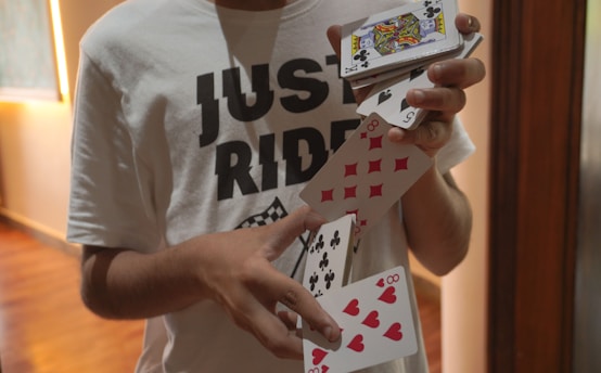 A person wearing a white T-shirt with the text 'JUST RIDE' is performing a card trick, spreading a deck of playing cards out in their hands. The background appears to be indoors with wooden flooring and a window letting in natural light.