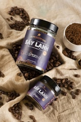 Two jars of freeze-dried instant coffee labeled 'Sấy Lạnh Aroma', surrounded by scattered coffee beans and a small bowl of coffee grounds, arranged on beige burlap fabric.