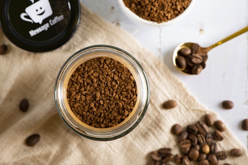 A top-down view of a jar filled with instant coffee granules placed on a light linen cloth. Nearby, a golden spoon holds coffee beans, and additional coffee beans are scattered around. The lid of the jar features a logo of a smiling coffee cup with the text 'Message Coffee'.