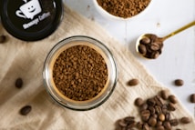 A top-down view of a jar filled with instant coffee granules placed on a light linen cloth. Nearby, a golden spoon holds coffee beans, and additional coffee beans are scattered around. The lid of the jar features a logo of a smiling coffee cup with the text 'Message Coffee'.