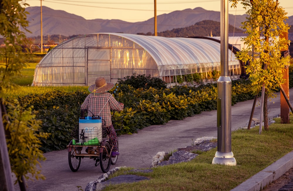 a person in a hat pushing a cart with plants and a greenhouse in the background