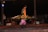 A performer in traditional attire is spinning in a brightly colored costume with intricate patterns. The event takes place in a nighttime setting, with several people seated in the background, watching the performance. The venue appears to be decorated with carpets and ambient lighting.