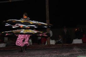 A performer spins in vibrant, layered skirts adorned with geometric patterns under dim lighting. Spectators are seated in the background, watching the performance on what appears to be a carpeted surface.