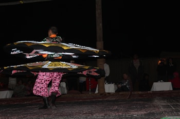 A performer spins in vibrant, layered skirts adorned with geometric patterns under dim lighting. Spectators are seated in the background, watching the performance on what appears to be a carpeted surface.