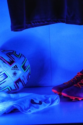 A soccer ball with the Adidas and Euro 2020 logos is positioned next to a pair of brightly colored soccer cleats and a folded jersey with the number 3. The scene is illuminated by a blue light, creating a vibrant and modern atmosphere.