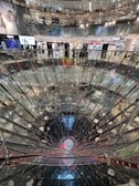 A large, multi-level shopping mall interior featuring a striking, deep cylindrical glass or mirror structure at the center. Surrounding the structure are various retail store fronts with bright, colorful signage and lighting. The mall's upper levels are visible with glass railings, creating a sense of openness and modern design.
