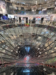 A large, multi-level shopping mall interior featuring a striking, deep cylindrical glass or mirror structure at the center. Surrounding the structure are various retail store fronts with bright, colorful signage and lighting. The mall's upper levels are visible with glass railings, creating a sense of openness and modern design.