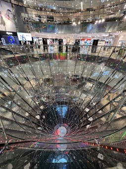 A large, multi-level shopping mall interior featuring a striking, deep cylindrical glass or mirror structure at the center. Surrounding the structure are various retail store fronts with bright, colorful signage and lighting. The mall's upper levels are visible with glass railings, creating a sense of openness and modern design.