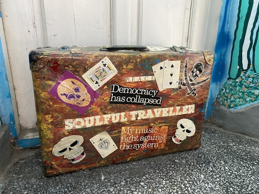 A worn-out suitcase is adorned with various punk-themed stickers and graffiti. Among the images are skulls, playing cards, and text like 'Democracy has collapsed,' 'Soulful Traveller,' and 'My music fight against the system.' The suitcase is set against a rustic background with a painted wall.