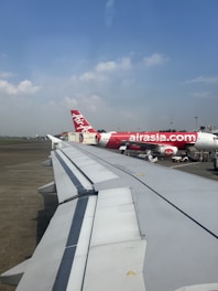 A view from an airplane window showing the wing of a plane on the runway. In the background, there is an AirAsia plane parked at the airport gate. The sky is clear with a few scattered clouds.