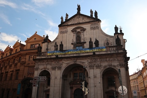 A historic building with detailed architectural features, including statues and ornate carvings. A large banner reading 'HANDS OFF UKRAINE, PUTIN!' is displayed prominently across the facade. The sky above is clear with some light clouds.