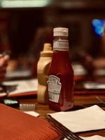 A bottle of Heinz tomato ketchup is placed on top of a wooden table, alongside a bottle of mustard. The setting appears to be a casual dining establishment with menus and cutlery visible. The background is blurred, highlighting the condiments in the foreground.