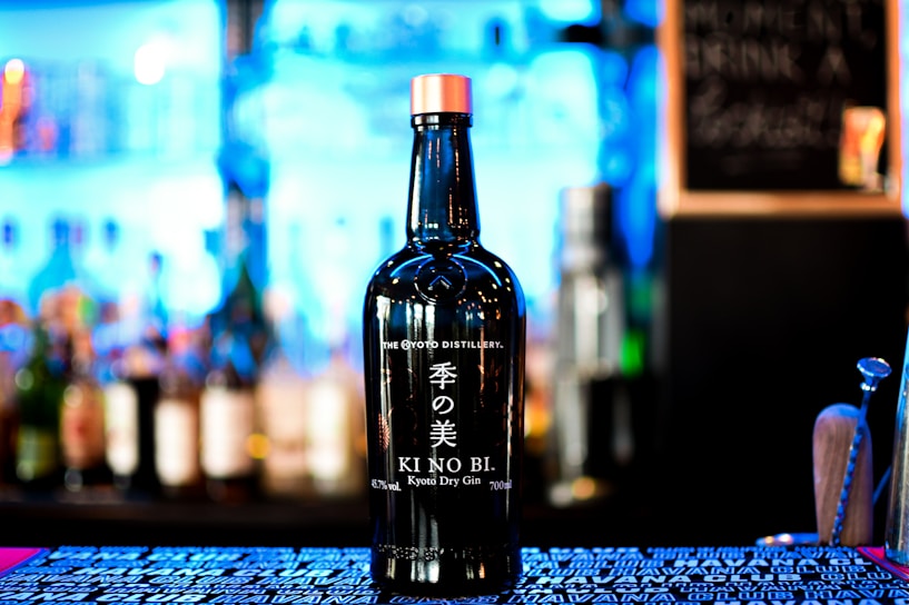 A bottle of KI NO BI Kyoto Dry Gin is prominently displayed against a blurred background of various bottles on shelves, suggesting a bar setting. The bottle has a sleek black design with white Japanese characters and English text.