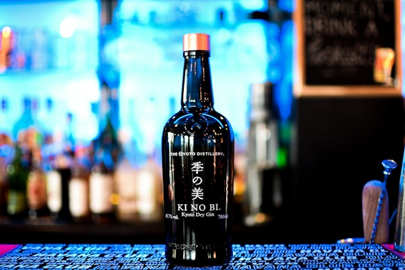 A bottle of KI NO BI Kyoto Dry Gin is prominently displayed against a blurred background of various bottles on shelves, suggesting a bar setting. The bottle has a sleek black design with white Japanese characters and English text.