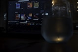 A dimly lit scene featuring a laptop with a music streaming service interface on the screen. Playlists and music recommendations are visible. A glass with water is positioned in front of the screen, capturing reflections and adding a subtle glow to the foreground.