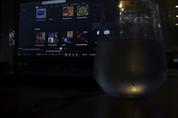 A dimly lit scene featuring a laptop with a music streaming service interface on the screen. Playlists and music recommendations are visible. A glass with water is positioned in front of the screen, capturing reflections and adding a subtle glow to the foreground.