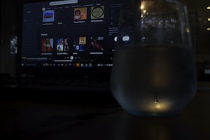 A dimly lit scene featuring a laptop with a music streaming service interface on the screen. Playlists and music recommendations are visible. A glass with water is positioned in front of the screen, capturing reflections and adding a subtle glow to the foreground.