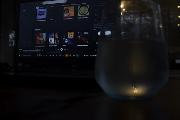 A dimly lit scene featuring a laptop with a music streaming service interface on the screen. Playlists and music recommendations are visible. A glass with water is positioned in front of the screen, capturing reflections and adding a subtle glow to the foreground.