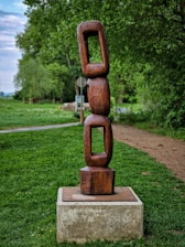 A wooden sculpture consisting of three interconnected, rectangular links stands on a concrete base in a lush green park setting. The background reveals trees and a path leading away from the sculpture.