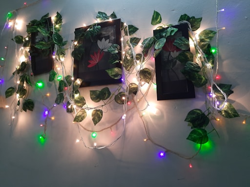 A wall is adorned with vine-like artificial green leaves intertwined with colorful string lights. The arrangement is centered around framed artworks featuring floral motifs, creating a lush, vibrant aesthetic.