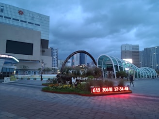 An urban area featuring a modern architectural landscape, dominated by a large building labeled 'Shinsegae.' The sky is overcast with dark gray clouds. In the foreground, there is a large digital display showing time and date information in red. A decorative garden with a basket-like structure containing plants and flowers adds a touch of nature. There is also a curved glass structure likely serving as an entrance or walkway.