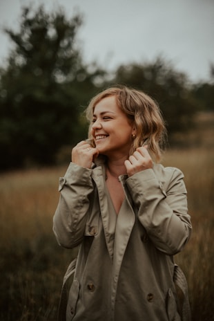 A person wearing a trench coat is standing outdoors in a field, smiling brightly. The background is blurred, with tall grass and trees visible, creating a natural, serene setting.