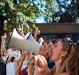 A group of people are gathered outdoors, with a woman prominently shouting into a megaphone. Surrounding her, several individuals have their fists raised or fingers pointing upward. Trees and a building are visible in the background.