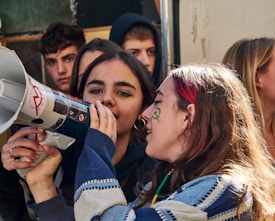A group of young people gathered together, with the focus on two individuals in the foreground. One person is holding and speaking into a megaphone, while the other appears to be listening or assisting. The background shows more people, all of whom seem to be engaged in the event. Some have face paint.