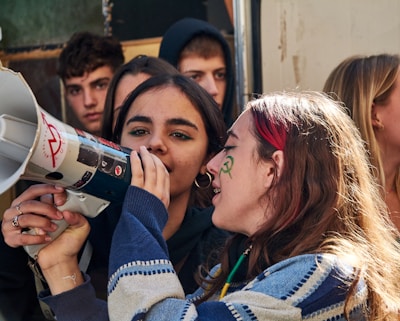 A group of young people gathered together, with the focus on two individuals in the foreground. One person is holding and speaking into a megaphone, while the other appears to be listening or assisting. The background shows more people, all of whom seem to be engaged in the event. Some have face paint.