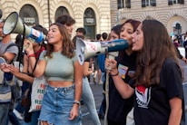 Several young people are participating in a street protest, using megaphones to amplify their voices. They appear animated and engaged, with some holding signs. The background features a building with arched windows and signs indicating shops.