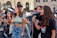 Several young people are participating in a street protest, using megaphones to amplify their voices. They appear animated and engaged, with some holding signs. The background features a building with arched windows and signs indicating shops.