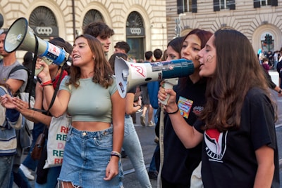 Several young people are participating in a street protest, using megaphones to amplify their voices. They appear animated and engaged, with some holding signs. The background features a building with arched windows and signs indicating shops.