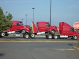Three red semi-truck cabs are stacked together in a parking lot. The trucks are positioned in a line, each cab is mounted on top of the one in front of it. The scene includes a stop sign to the right and a patch of greenery on the left.