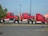 Three red semi-truck cabs are stacked together in a parking lot. The trucks are positioned in a line, each cab is mounted on top of the one in front of it. The scene includes a stop sign to the right and a patch of greenery on the left.
