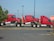 Three red semi-truck cabs are stacked together in a parking lot. The trucks are positioned in a line, each cab is mounted on top of the one in front of it. The scene includes a stop sign to the right and a patch of greenery on the left.