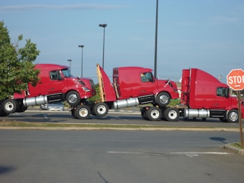 Three red semi-truck cabs are stacked together in a parking lot. The trucks are positioned in a line, each cab is mounted on top of the one in front of it. The scene includes a stop sign to the right and a patch of greenery on the left.