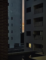 Tall, modern apartment buildings with several windows and balconies frame a narrow view of a colorful sunset in the distance. The lighting is low, suggesting evening or early night, with some lights visible inside the apartments.