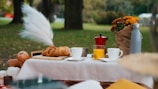 A picnic setup on a table covered with a white tablecloth. The arrangement includes croissants on a wooden board, a red coffee maker, a glass of orange juice, and several cups. A gray thermos and a bouquet of orange flowers wrapped in brown paper are also present. The setting appears to be in a park with green grass and tall trees in the background.